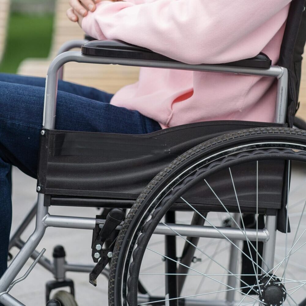 A young adult in a pink hoodie sits in a wheelchair outdoors, showcasing inclusivity and accessibility.