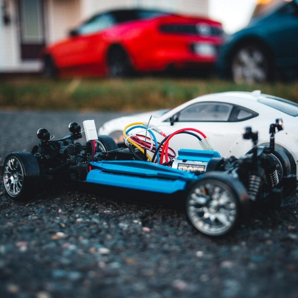 Close-up of a remote control car chassis with colorful wires on asphalt, with blurred cars in background.